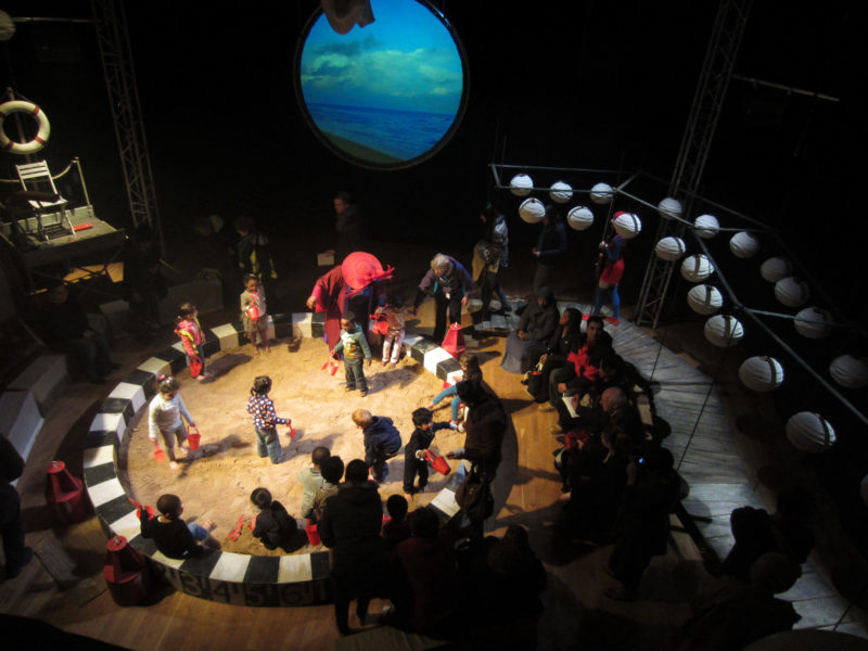 A bird’s eye view of the audience arriving for an under-fives show set at the seaside. Children with buckets and spades are being welcomed to the A bird’s eye view of the audience arriving for an under-fives show set at the seaside. Children with buckets and spades are being welcomed to the sandpit by a performer in a twenties beach outfit. On one side, there is a wooden jetty and on the other, the raised platform for the musician-come-lifeguard. On the back wall, there is a screen showing video of the sea and sky on an empty beach.