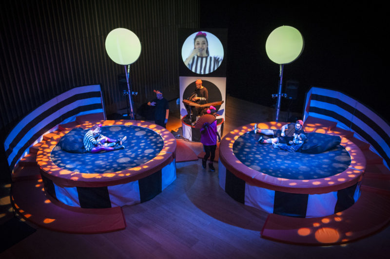 There are two trampolines side by side. A child and a trampolinist are sitting on each one with a musician playing in between and video screens above them.